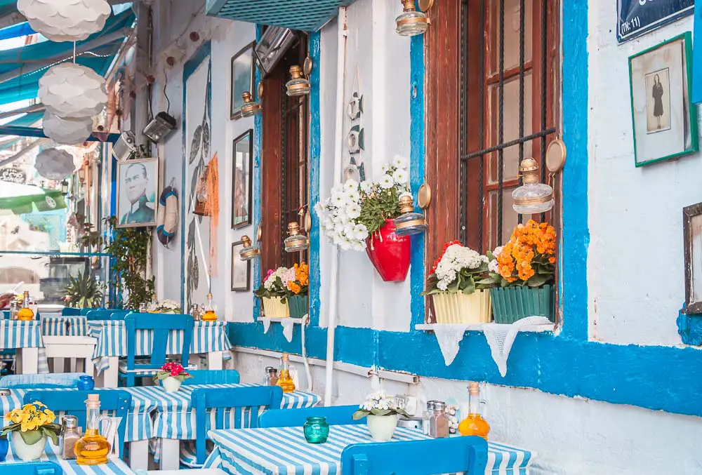 Eine der buten Hausfassaden in Alacati mit blauen Fensterläden, Blumen, blauen Sesseln, weißen Tischen und weißen Wänden. 